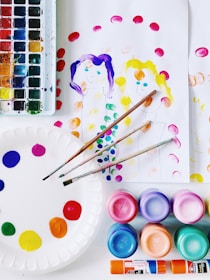 A collection of art supplies is displayed, including a watercolor palette with various colors, brushes, and several containers of vibrant paints. There's a paper plate used as a palette with dabs of paint, along with sheets of paper featuring simple, colorful paintings of two people. The scene is bright and colorful, suggesting an art or craft activity.