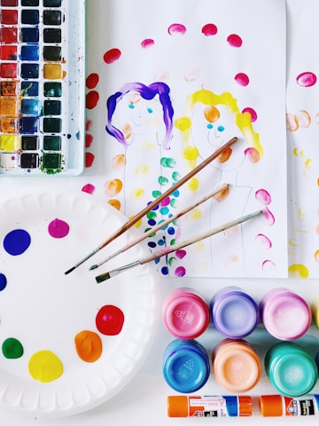 A collection of art supplies is displayed, including a watercolor palette with various colors, brushes, and several containers of vibrant paints. There's a paper plate used as a palette with dabs of paint, along with sheets of paper featuring simple, colorful paintings of two people. The scene is bright and colorful, suggesting an art or craft activity.