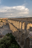 A long, ancient stone aqueduct stretches across a cityscape under a partly cloudy sky, casting shadows on the ground below. The structure features multiple arches and is surrounded by buildings and trees. People can be seen walking near the aqueduct, giving a sense of scale to the large construction.