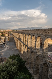 A long, ancient stone aqueduct stretches across a cityscape under a partly cloudy sky, casting shadows on the ground below. The structure features multiple arches and is surrounded by buildings and trees. People can be seen walking near the aqueduct, giving a sense of scale to the large construction.
