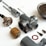 A collection of disassembled metallic mechanical parts arranged on a white surface. The parts include various cylindrical components, screws, and a rusty circular gasket. The components vary in size and shape, with some showing signs of wear.