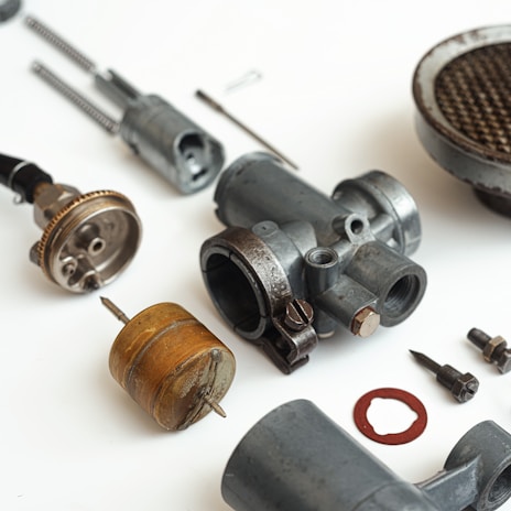 A collection of disassembled metallic mechanical parts arranged on a white surface. The parts include various cylindrical components, screws, and a rusty circular gasket. The components vary in size and shape, with some showing signs of wear.