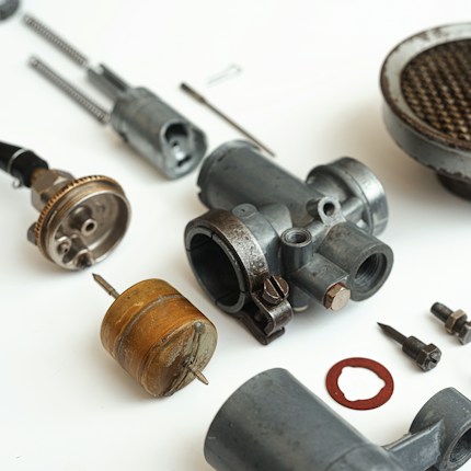 A collection of disassembled metallic mechanical parts arranged on a white surface. The parts include various cylindrical components, screws, and a rusty circular gasket. The components vary in size and shape, with some showing signs of wear.