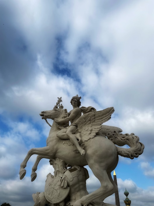 A statue depicting a mythological figure riding a winged horse against a backdrop of a partly cloudy sky. The sculpture is detailed with intricate textures, capturing both movement and grace. The horse is rearing, and the rider appears poised and commanding, embodying a sense of classical elegance.