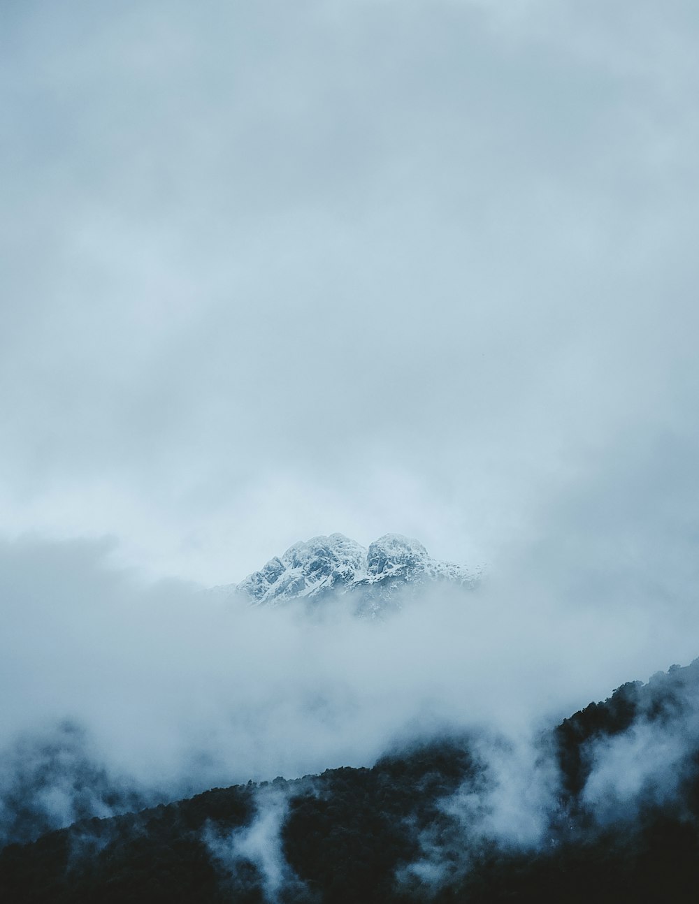 a mountain covered in clouds with a snow capped mountain in the distance