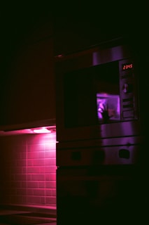 A dimly lit kitchen features a tiled backsplash illuminated by a purple light with a microwave oven above the countertop. The microwave's digital display reads 23:45, and the interior light casts an eerie silhouette of a hand on the microwave door.