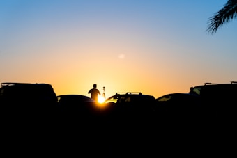 The silhouette of a person with a tripod stands against a vibrant sunset, with various vehicles around and a palm tree visible on the right. The sky transitions from deep blue at the top to golden near the horizon.