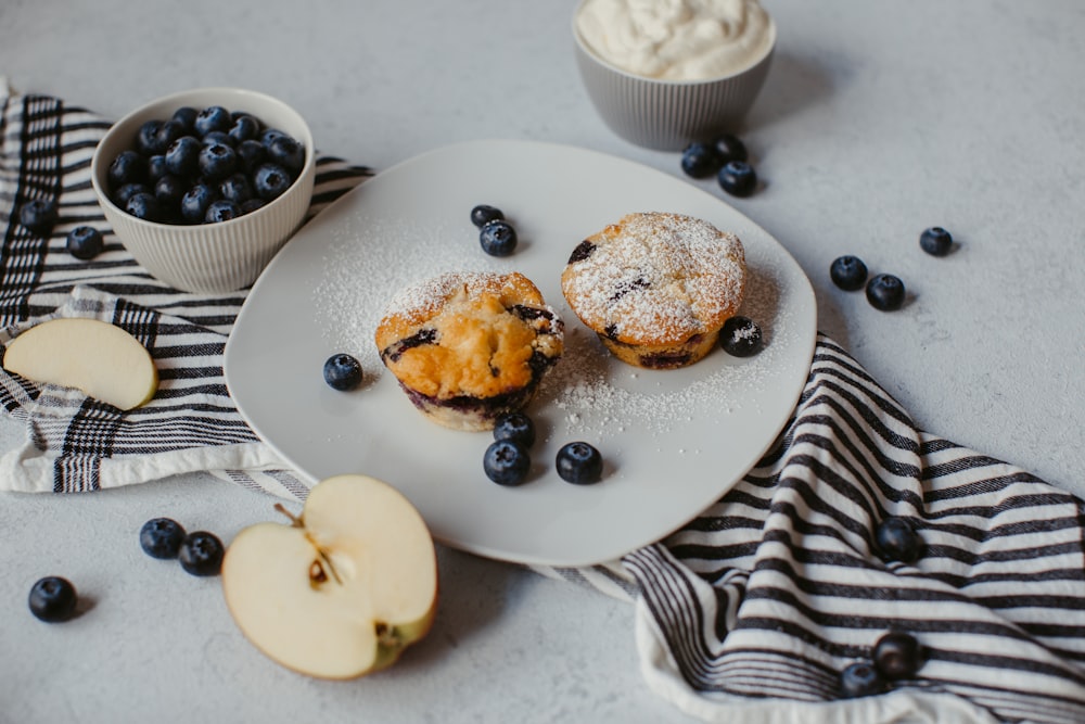 a white plate topped with blueberry muffins next to a bowl of blue