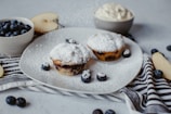Two blueberry muffins sit on a white plate, dusted with powdered sugar. Beside them, fresh blueberries are scattered. A striped cloth adds texture to the composition. A bowl of creamy topping and a cut apple are placed in the background, adding to the rustic aesthetic.