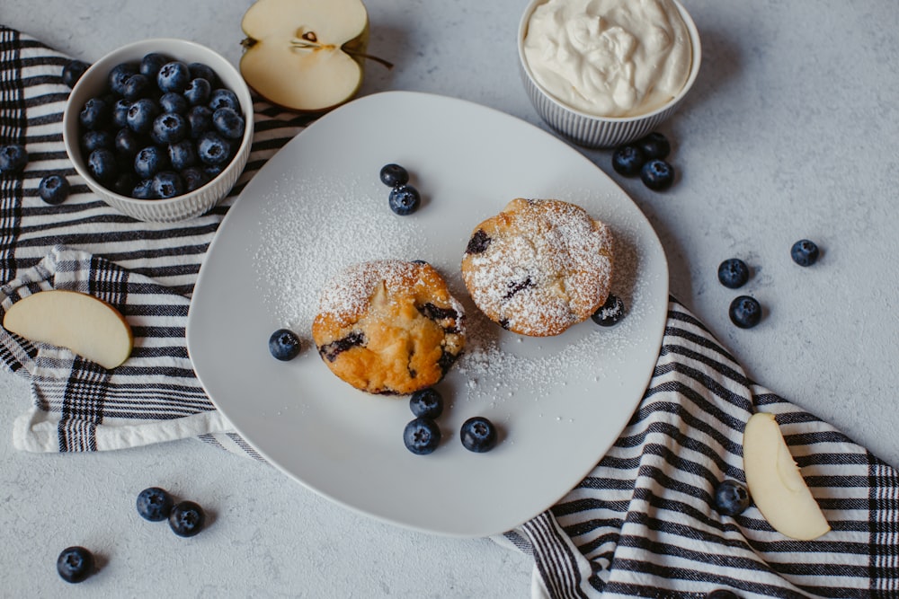 a white plate topped with blueberry muffins next to a bowl of blue