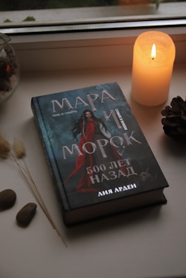 A hardcover book lies on a windowsill next to a lit candle, stones, dried grass, and a small decorative item. The book cover features a dark theme with text in a non-English language and an illustration of a woman in red.