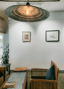 A cozy indoor seating area featuring wooden furniture including chairs and a table. Above the seating area hangs a large, woven rattan light fixture with an exposed bulb. The white wall behind the seating area displays two framed artworks. One is a portrait and the other is a landscape sketch. There is also a potted plant on the left side of the room. The decor has a natural and minimalist aesthetic.