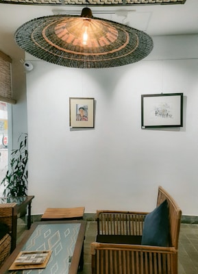 A cozy indoor seating area featuring wooden furniture including chairs and a table. Above the seating area hangs a large, woven rattan light fixture with an exposed bulb. The white wall behind the seating area displays two framed artworks. One is a portrait and the other is a landscape sketch. There is also a potted plant on the left side of the room. The decor has a natural and minimalist aesthetic.