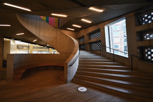 A modern architectural interior featuring a large spiral staircase with concrete and wooden elements. Soft lighting from ceiling fixtures highlights the geometric design, and a large window to the right offers a glimpse of an exterior structure.