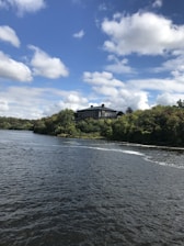 A serene landscape with a large, modern house on a lush, green hillside. The house overlooks a calm river, with trees surrounding the area. The sky is bright blue, filled with scattered white clouds.