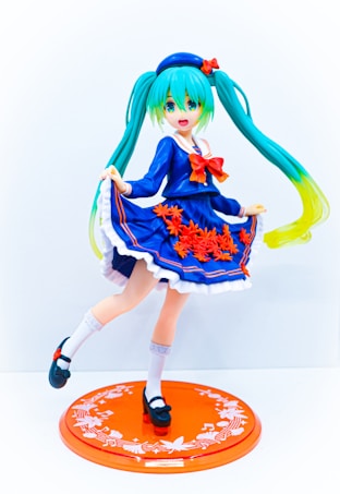 A figure of an anime character with teal twin-tails and bright green eyes, dressed in a navy blue dress adorned with red flowers and a red bow. The character stands on one leg with the other slightly raised, showcasing white socks and black shoes. The dress has white frilled hem and the figure is mounted on an orange circular base with white decorative patterns.