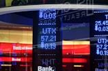 A digital display board showcasing numerical data with bright blue LED numbers. A glass reflection reveals partial text and a logo related to a bank. The background features vibrant red and white colors with the visible text 'Bank of America'.