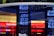 A digital display board showcasing numerical data with bright blue LED numbers. A glass reflection reveals partial text and a logo related to a bank. The background features vibrant red and white colors with the visible text 'Bank of America'.