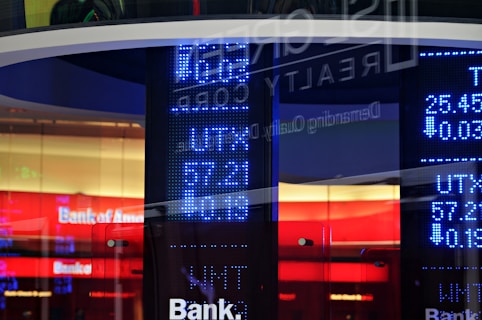 A digital display board showcasing numerical data with bright blue LED numbers. A glass reflection reveals partial text and a logo related to a bank. The background features vibrant red and white colors with the visible text 'Bank of America'.