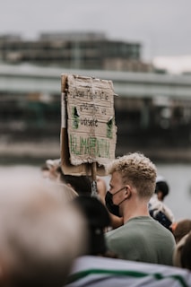 A person with curly blonde hair, wearing a black face mask and green shirt, stands in a crowd holding a cardboard protest sign with German text. The background is blurred, suggesting an outdoor urban setting.