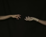 Two hands reaching towards each other against a dark background. The left hand is bare, while the right hand wears a glove. Both hands are outstretched, with fingers partially curled and almost touching.