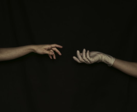 Two hands reaching towards each other against a dark background. The left hand is bare, while the right hand wears a glove. Both hands are outstretched, with fingers partially curled and almost touching.