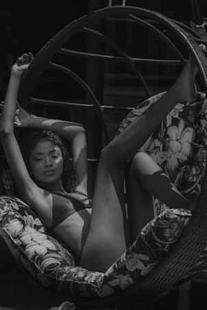 A person is lounging comfortably on a circular wicker chair with floral-patterned cushions, wearing swimwear and appearing relaxed with an arm and leg positioned upwards.