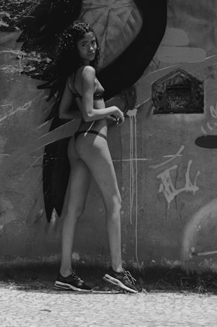A woman stands against a graffiti-covered wall, wearing a bikini and sneakers. She has curly hair and is turned slightly towards the camera with a subtle smile. The artwork on the wall features abstract shapes and a stylized eye.