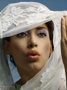 A woman with styled makeup and glossy lips is partially covered by a decorative lace veil. The veil features intricate floral embroidery. Her eyes are accentuated with eyeshadow, and she wears a pair of small earrings. The background is a soft blue, complementing the overall aesthetic.