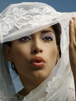 A woman with styled makeup and glossy lips is partially covered by a decorative lace veil. The veil features intricate floral embroidery. Her eyes are accentuated with eyeshadow, and she wears a pair of small earrings. The background is a soft blue, complementing the overall aesthetic.