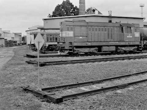 A black and white photograph featuring a railway yard with a locomotive positioned on the tracks. The background includes industrial buildings and a chimney, along with a small signal sign near the tracks. The area appears to be part of a larger, possibly historical, rail facility.