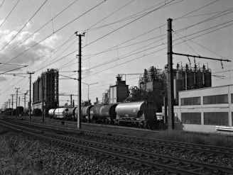 Railway tracks run parallel to a large industrial facility with multiple tanks and piping structures. Several cylindrical train cars are positioned on the tracks, and power lines are visible above. The scene is in black and white, giving it a timeless and industrial character.