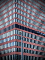A modern high-rise building with alternating red and blue horizontal stripes. Numerous rows of windows create a repetitive pattern across the structure. A lift or maintenance platform is attached to the facade, suggesting building maintenance or window cleaning activities. A tall streetlight is positioned in the foreground.