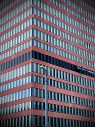 A modern high-rise building with alternating red and blue horizontal stripes. Numerous rows of windows create a repetitive pattern across the structure. A lift or maintenance platform is attached to the facade, suggesting building maintenance or window cleaning activities. A tall streetlight is positioned in the foreground.
