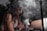 A woman with long, wavy hair is working on a tattoo. She wears a white top and gloves, and appears focused on the task as she leans over a person's leg. Smoke or mist partially obscures the scene, adding a mysterious atmosphere.