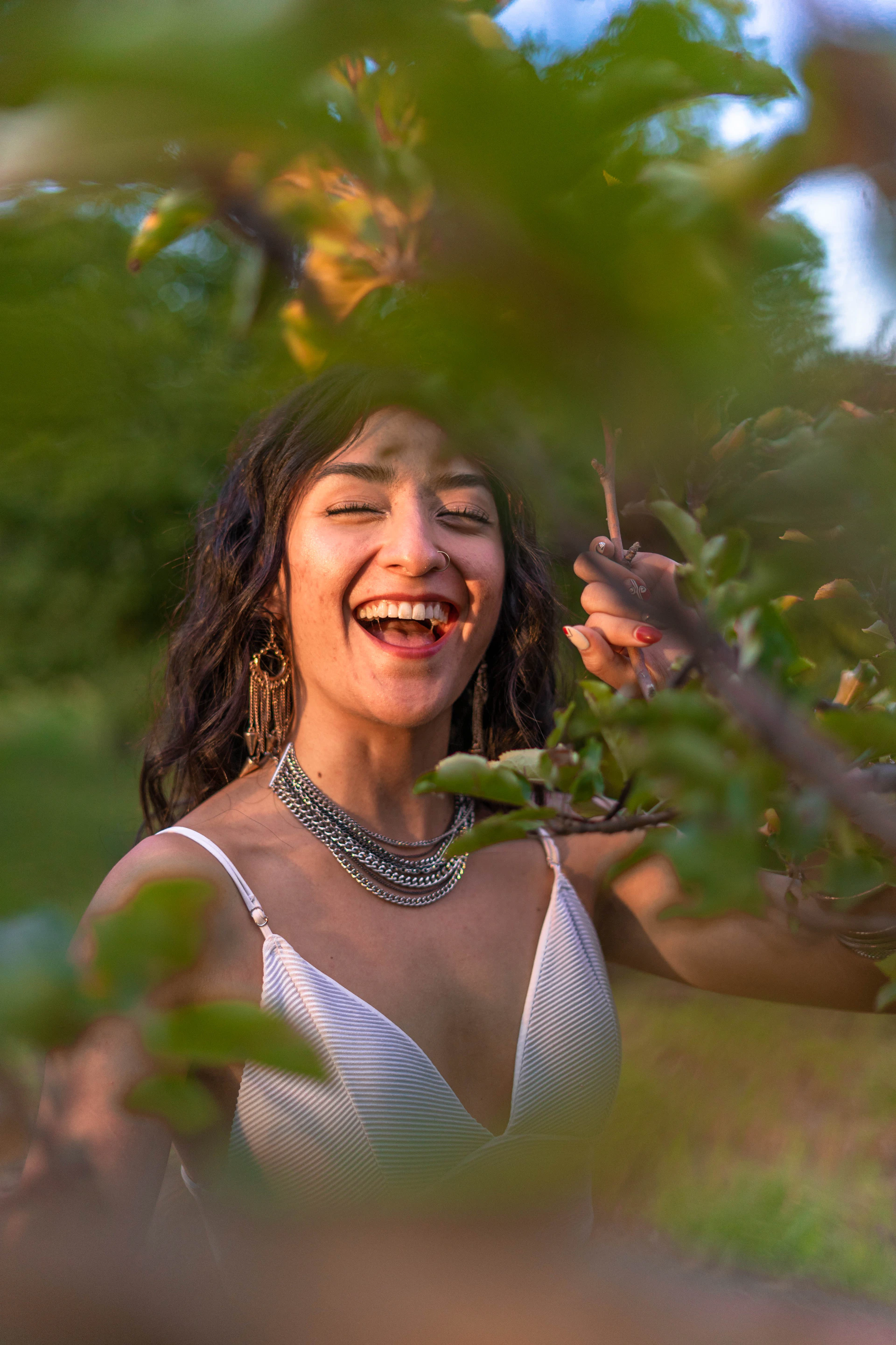 A candid shot of a woman laughing outdoors, wearing a light linen outfit, with natural greenery softly blurred in the background.
