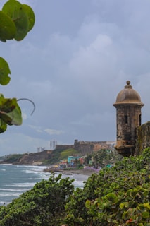 A coastal landscape featuring a historical stone watchtower near the shore. The scene includes lush green foliage in the foreground and a stretch of beach with gentle waves. In the background, colorful buildings are situated near the water's edge, with larger stone fortifications and modern buildings further away.