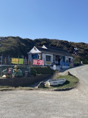A small, white building with a dark roof sits by the roadside, surrounded by grassy hills. The building has colorful signs and banners displayed outside, and a variety of beach toys or inflatables are visible. Nearby, a small rowboat is placed on the grass. The sky is clear and blue, suggesting a sunny day.