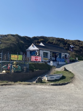 A small, white building with a dark roof sits by the roadside, surrounded by grassy hills. The building has colorful signs and banners displayed outside, and a variety of beach toys or inflatables are visible. Nearby, a small rowboat is placed on the grass. The sky is clear and blue, suggesting a sunny day.