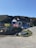 A small, white building with a dark roof sits by the roadside, surrounded by grassy hills. The building has colorful signs and banners displayed outside, and a variety of beach toys or inflatables are visible. Nearby, a small rowboat is placed on the grass. The sky is clear and blue, suggesting a sunny day.