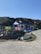 A small, white building with a dark roof sits by the roadside, surrounded by grassy hills. The building has colorful signs and banners displayed outside, and a variety of beach toys or inflatables are visible. Nearby, a small rowboat is placed on the grass. The sky is clear and blue, suggesting a sunny day.