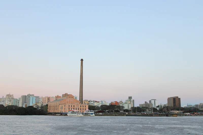 Pôr do sol sobre o rio Guaíba em Porto Alegre, com a água do rio em primeiro plano e a silhueta dos prédios do centro na outra margem