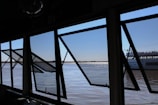 Several large open windows frame a view of a calm river with a distant shoreline. Through the windows, part of a boat with the name 'Porto Alegre' is visible. The sky is clear, and the water's surface reflects sunlight, creating a shimmering effect.