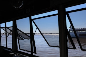 Several large open windows frame a view of a calm river with a distant shoreline. Through the windows, part of a boat with the name 'Porto Alegre' is visible. The sky is clear, and the water's surface reflects sunlight, creating a shimmering effect.