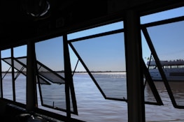 Several large open windows frame a view of a calm river with a distant shoreline. Through the windows, part of a boat with the name 'Porto Alegre' is visible. The sky is clear, and the water's surface reflects sunlight, creating a shimmering effect.