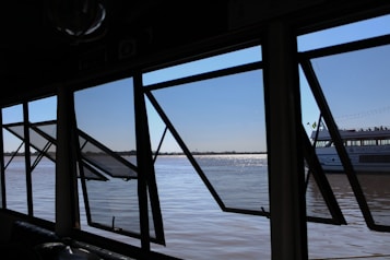 Several large open windows frame a view of a calm river with a distant shoreline. Through the windows, part of a boat with the name 'Porto Alegre' is visible. The sky is clear, and the water's surface reflects sunlight, creating a shimmering effect.