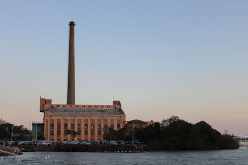 A large industrial building with a tall smokestack stands on the waterfront. The structure is surrounded by parked cars, and a dense area of trees or bushes is visible nearby. The sky is clear, transitioning from a light blue at the top to a soft pink hue near the horizon.