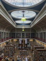 A large, classic library with tall shelves filled with books. The roof is adorned with an intricate glass dome ceiling allowing natural light to illuminate the space. There are several tables and chairs arranged for reading or study, and statues are placed between the shelves. The atmosphere is quiet and scholarly.
