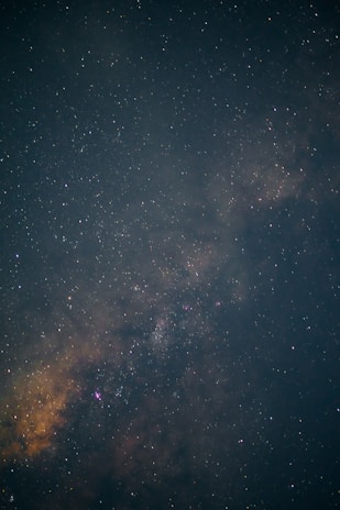 A vast view of the night sky filled with countless stars and subtle nebulae formations in varying shades of dark blue and orange. The stars create a speckled pattern across the entire image, revealing the depth and expanse of the universe.