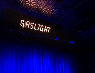 A dimly lit stage with the word 'GASLIGHT' in glowing white neon letters above. The backdrop features dark blue curtains, and the ceiling displays an ornate pattern. The overall atmosphere suggests a vintage or intimate performance space.